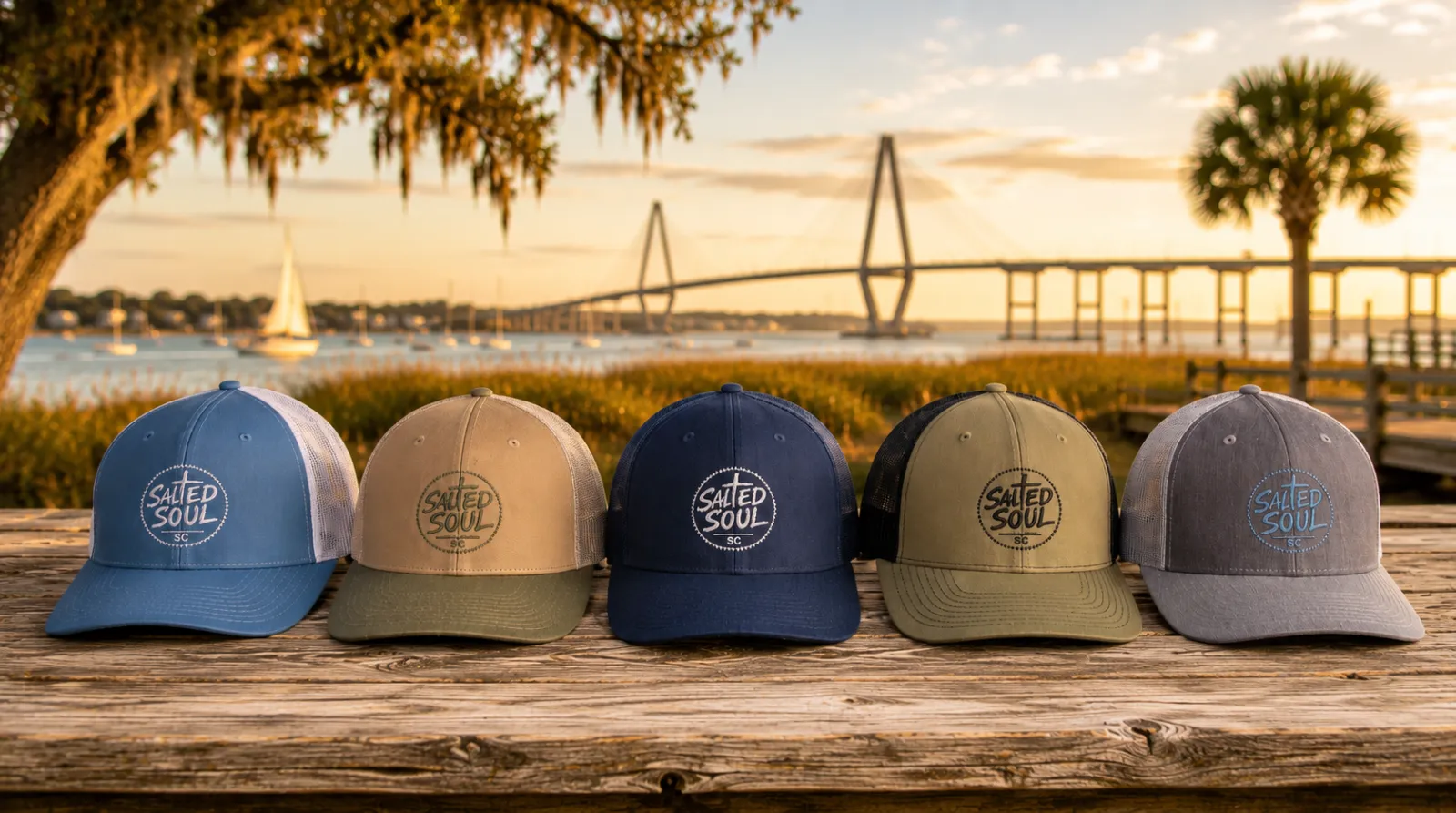 Five Salted Soul trucker hats lined up on a dock table with the Ravenel Bridge and Charleston harbor behind them